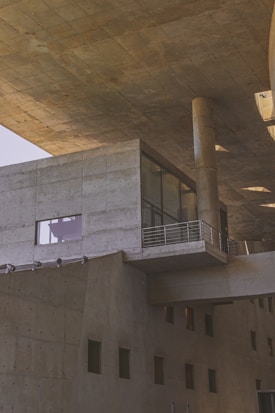 A modern architectural structure featuring large concrete surfaces, angular lines, and a robust design. The building includes various geometric shapes, such as rectangular windows and cylindrical columns, providing a sense of solidity and industrial aesthetic. The lighting casts subtle shadows on the textured concrete, enhancing the architectural details.