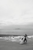 A person standing on a beach near the shoreline, casting a large fishing net into the ocean. Waves are gently rolling onto the sand, and a small island is visible on the horizon under an overcast sky.