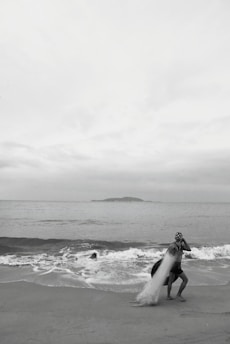 A person standing on a beach near the shoreline, casting a large fishing net into the ocean. Waves are gently rolling onto the sand, and a small island is visible on the horizon under an overcast sky.