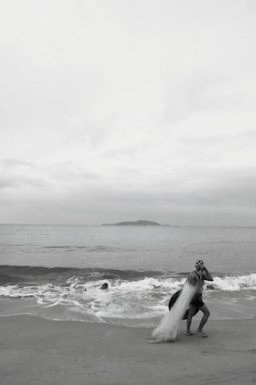 A person standing on a beach near the shoreline, casting a large fishing net into the ocean. Waves are gently rolling onto the sand, and a small island is visible on the horizon under an overcast sky.
