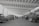 A spacious indoor garage or workshop features rows of vintage cars, including several Volkswagen Beetles. The vehicles are arranged neatly, some undergoing maintenance or being worked on by mechanics. Large windows line the walls, allowing natural light to illuminate the expansive area. Two men stand conversing in the center, seemingly discussing the cars or repairs.