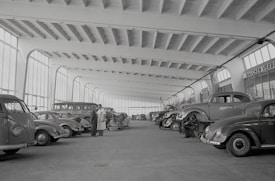 A spacious indoor garage or workshop features rows of vintage cars, including several Volkswagen Beetles. The vehicles are arranged neatly, some undergoing maintenance or being worked on by mechanics. Large windows line the walls, allowing natural light to illuminate the expansive area. Two men stand conversing in the center, seemingly discussing the cars or repairs.