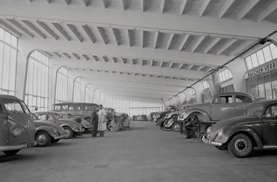 A spacious indoor garage or workshop features rows of vintage cars, including several Volkswagen Beetles. The vehicles are arranged neatly, some undergoing maintenance or being worked on by mechanics. Large windows line the walls, allowing natural light to illuminate the expansive area. Two men stand conversing in the center, seemingly discussing the cars or repairs.