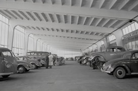 A spacious indoor garage or workshop features rows of vintage cars, including several Volkswagen Beetles. The vehicles are arranged neatly, some undergoing maintenance or being worked on by mechanics. Large windows line the walls, allowing natural light to illuminate the expansive area. Two men stand conversing in the center, seemingly discussing the cars or repairs.