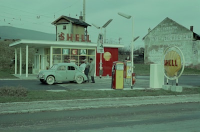 A retro Shell gas station features an old-fashioned car being refueled by two attendants. The station has distinct red and yellow fuel pumps, a prominent Shell sign, and a vintage building design. The surrounding area includes another Shell sign with the iconic shell logo and a weathered building in the background.