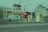 A retro Shell gas station features an old-fashioned car being refueled by two attendants. The station has distinct red and yellow fuel pumps, a prominent Shell sign, and a vintage building design. The surrounding area includes another Shell sign with the iconic shell logo and a weathered building in the background.