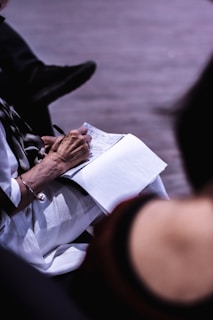 An individual is writing notes on a notepad, using a pen. The person's attire includes a light-colored garment with a bracelet. The focus is on the hands and notepad, while other details are softly blurred in the background.