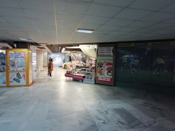 An indoor market area features a variety of small retail displays, including a food stall with fresh produce and a prominently displayed advertisement. The floor is polished and tiled, and there is ample artificial lighting from the ceiling fixtures. A person in warm clothing walks through the space, adding a sense of movement.
