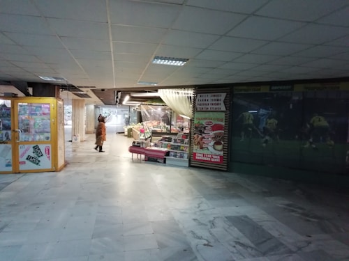 An indoor market area features a variety of small retail displays, including a food stall with fresh produce and a prominently displayed advertisement. The floor is polished and tiled, and there is ample artificial lighting from the ceiling fixtures. A person in warm clothing walks through the space, adding a sense of movement.