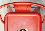 A top-down view of a red metal chair with curved armrests and a solid back. The chair has a rectangular seat with a cut-out detail.