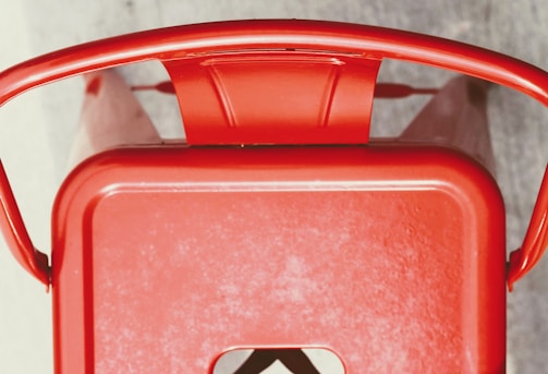 A top-down view of a red metal chair with curved armrests and a solid back. The chair has a rectangular seat with a cut-out detail.