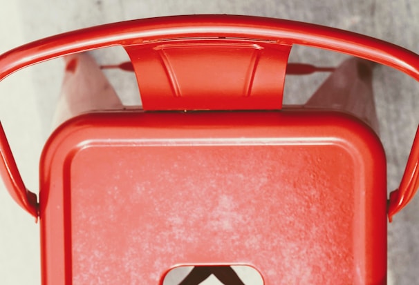 A top-down view of a red metal chair with curved armrests and a solid back. The chair has a rectangular seat with a cut-out detail.