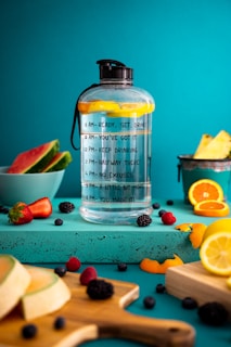 A large water bottle with motivational time markers sits on a turquoise surface surrounded by fresh fruits including slices of watermelon, strawberries, orange slices, and various berries. The background is a bright teal color that enhances the vibrancy of the fruit colors.