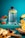 A large water bottle with motivational time markers sits on a turquoise surface surrounded by fresh fruits including slices of watermelon, strawberries, orange slices, and various berries. The background is a bright teal color that enhances the vibrancy of the fruit colors.