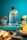 A large water bottle with motivational time markers sits on a turquoise surface surrounded by fresh fruits including slices of watermelon, strawberries, orange slices, and various berries. The background is a bright teal color that enhances the vibrancy of the fruit colors.