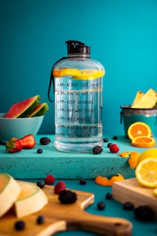 A large water bottle with motivational time markers sits on a turquoise surface surrounded by fresh fruits including slices of watermelon, strawberries, orange slices, and various berries. The background is a bright teal color that enhances the vibrancy of the fruit colors.