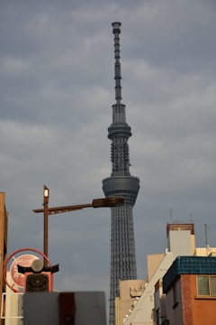 A towering communications and observation structure rises prominently against a cloudy sky. Surrounding buildings and a street sign are visible in the foreground, suggesting an urban setting.