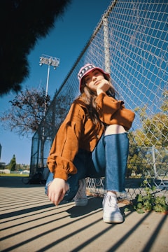 woman in brown coat and blue denim jeans sitting on gray metal fence during daytime