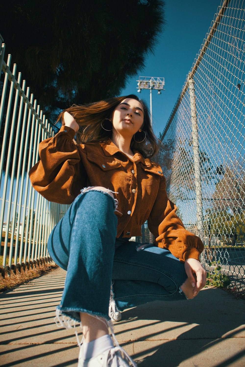 woman in brown coat and blue denim jeans sitting on brown wooden fence during daytime