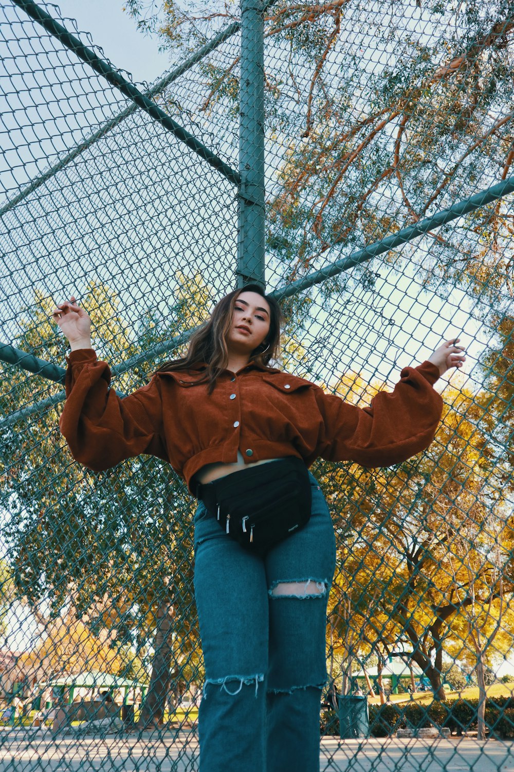 woman in brown jacket and blue denim jeans standing beside gray metal fence during daytime