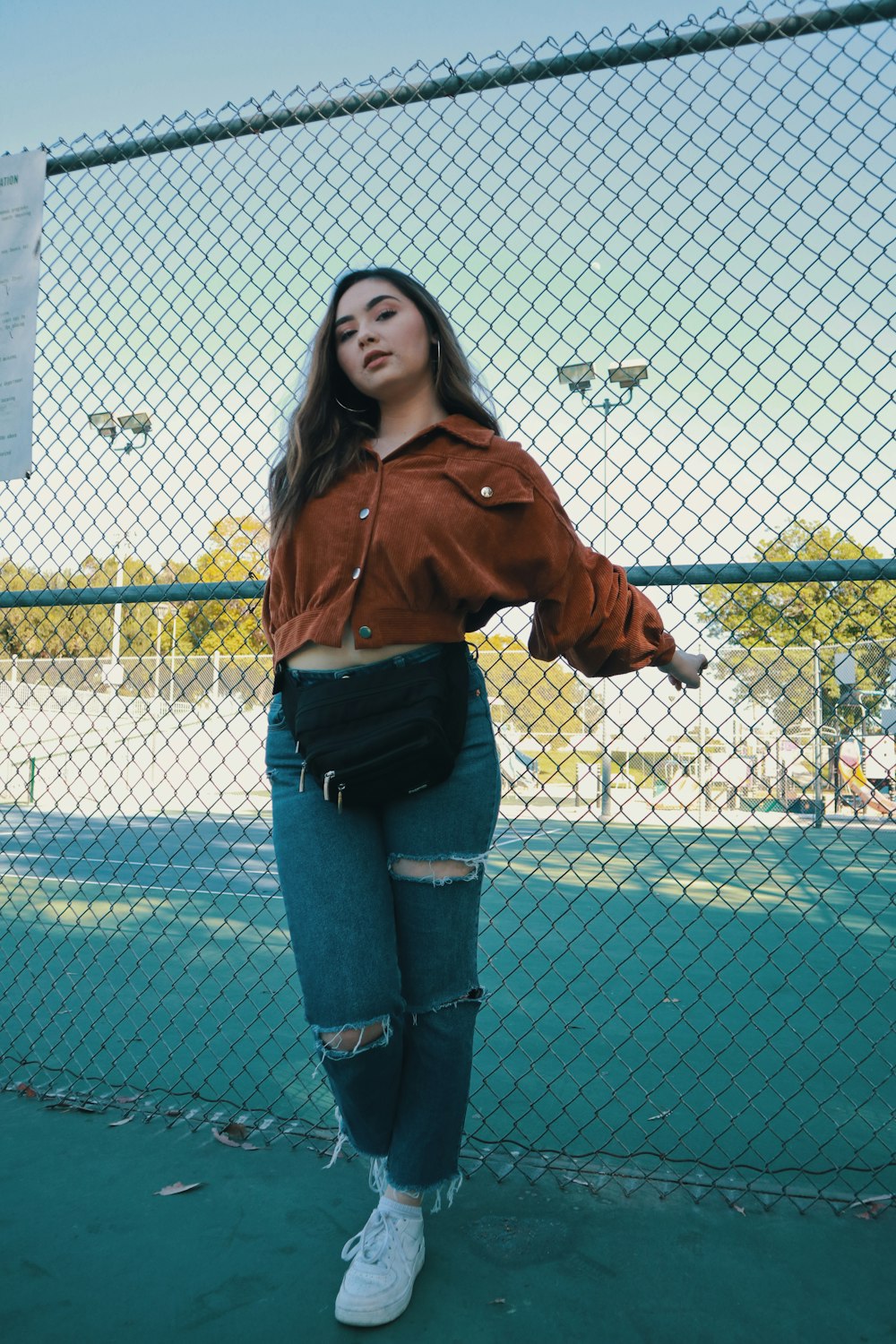 woman in brown jacket and blue denim jeans standing beside chain link fence