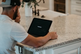 A person sits at a kitchen counter with their hand on their chin, looking at an open tablet device displaying the 'YouVersion' app. The countertop is made of light-colored granite, and there is a white vase with plants in the background.