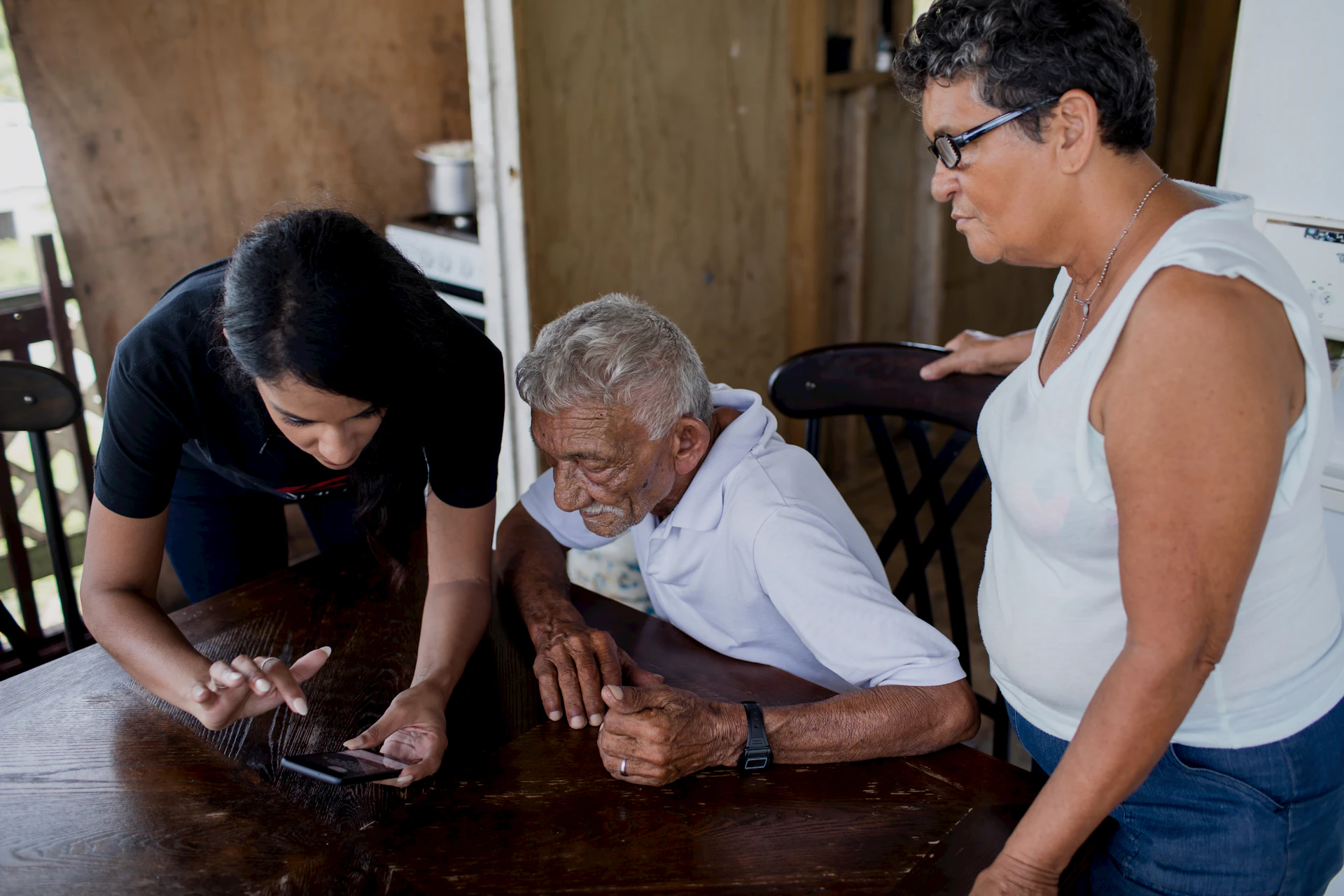A caregiver helping an older man look at a phone while a family member looks on at home