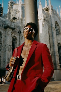 A person stands confidently in front of a grand, gothic-style cathedral. The individual is wearing a striking red suit with no shirt underneath, accessorized with bold jewelry and large white sunglasses. The sun casts a warm glow, highlighting both the person's attire and the intricate architecture of the building, which features spires and detailed carvings.