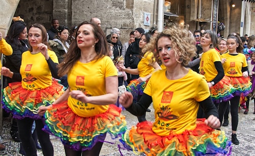 A group of women participate in a street parade, wearing bright yellow shirts with printed designs and matching rainbow-colored tutus. The scene is lively, with a small crowd observing in the background, and confetti scattered on the ground.
