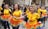 A group of women participate in a street parade, wearing bright yellow shirts with printed designs and matching rainbow-colored tutus. The scene is lively, with a small crowd observing in the background, and confetti scattered on the ground.