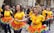 A group of women participate in a street parade, wearing bright yellow shirts with printed designs and matching rainbow-colored tutus. The scene is lively, with a small crowd observing in the background, and confetti scattered on the ground.