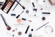 A collection of makeup products is artfully arranged on a white surface. The assortment includes eyeshadow palettes, lipsticks, brushes, mascara, and various containers of powder and cream cosmetics. The items display brand names like Maxeybrow, Maxeylash, and BareMinerals.