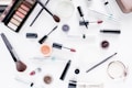 A collection of makeup products is artfully arranged on a white surface. The assortment includes eyeshadow palettes, lipsticks, brushes, mascara, and various containers of powder and cream cosmetics. The items display brand names like Maxeybrow, Maxeylash, and BareMinerals.