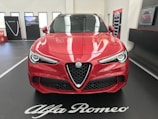 A red Alfa Romeo car is displayed indoors on a black platform with white lettering. The car's sleek design features prominent headlights and a distinctive grille. In the background, promotional panels and a screen are visible, enhancing the showroom setting.