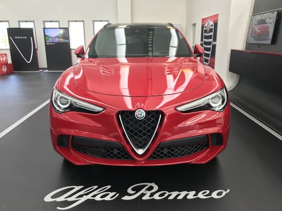 A red Alfa Romeo car is displayed indoors on a black platform with white lettering. The car's sleek design features prominent headlights and a distinctive grille. In the background, promotional panels and a screen are visible, enhancing the showroom setting.