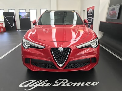 A red Alfa Romeo car is displayed indoors on a black platform with white lettering. The car's sleek design features prominent headlights and a distinctive grille. In the background, promotional panels and a screen are visible, enhancing the showroom setting.
