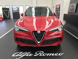 A red Alfa Romeo car is displayed indoors on a black platform with white lettering. The car's sleek design features prominent headlights and a distinctive grille. In the background, promotional panels and a screen are visible, enhancing the showroom setting.