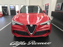 A red Alfa Romeo car is displayed indoors on a black platform with white lettering. The car's sleek design features prominent headlights and a distinctive grille. In the background, promotional panels and a screen are visible, enhancing the showroom setting.