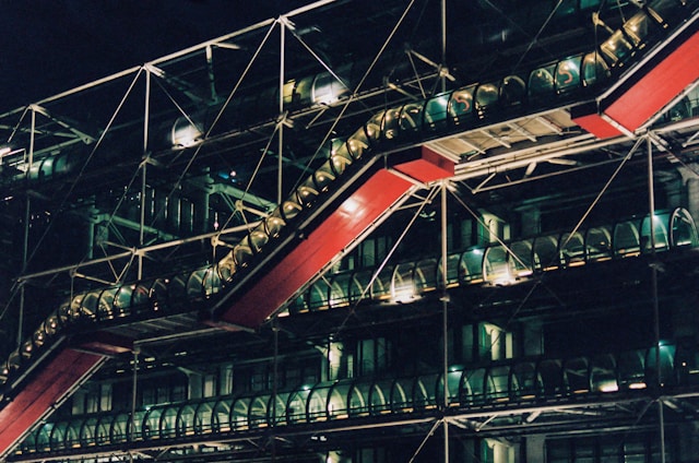 A complex architectural structure featuring exposed industrial components, including steel beams and transparent tubing. A prominent red external escalator contrasts with the dark framework. The building is illuminated subtly, highlighting its modern design against the night sky.