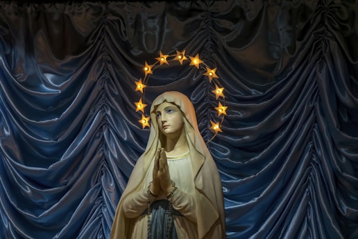 A statue of a woman in a cloak standing with hands clasped in prayer, surrounded by a halo of glowing stars. The backdrop is a luxurious blue curtain with a silky texture.