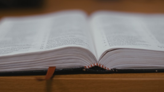An open book resting on a wooden surface, with visible text on its pages. A red bookmark ribbon can be seen hanging over the edge of the book.