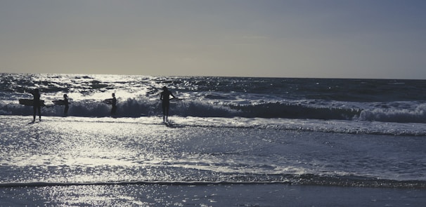 Silhouetted surfers stand with their boards at the edge of the ocean, with waves crashing around them and the sun reflecting off the water's surface.