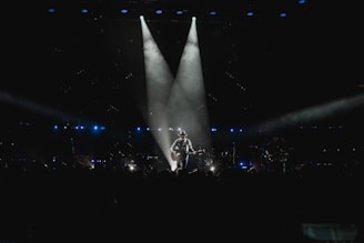 A musician stands on stage holding a guitar, illuminated by a spotlight against a dark background. The scene is surrounded by subtle blue lighting and patterns of small lights, suggesting an intimate atmosphere. The silhouette of the performer creates a dramatic focal point in the center.