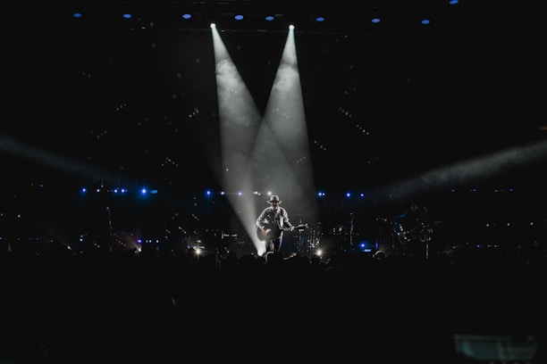 A musician stands on stage holding a guitar, illuminated by a spotlight against a dark background. The scene is surrounded by subtle blue lighting and patterns of small lights, suggesting an intimate atmosphere. The silhouette of the performer creates a dramatic focal point in the center.