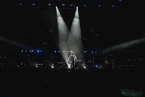 A musician stands on stage holding a guitar, illuminated by a spotlight against a dark background. The scene is surrounded by subtle blue lighting and patterns of small lights, suggesting an intimate atmosphere. The silhouette of the performer creates a dramatic focal point in the center.