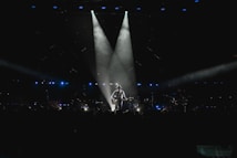 A musician stands on stage holding a guitar, illuminated by a spotlight against a dark background. The scene is surrounded by subtle blue lighting and patterns of small lights, suggesting an intimate atmosphere. The silhouette of the performer creates a dramatic focal point in the center.