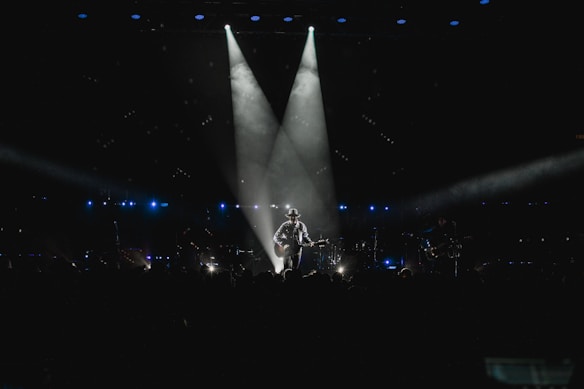 A musician stands on stage holding a guitar, illuminated by a spotlight against a dark background. The scene is surrounded by subtle blue lighting and patterns of small lights, suggesting an intimate atmosphere. The silhouette of the performer creates a dramatic focal point in the center.