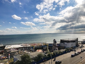 A seaside view with a coastline amusement park featuring various rides and attractions. The ocean stretches out under a partly cloudy sky, while people and cars can be seen on a road running parallel to the beach.