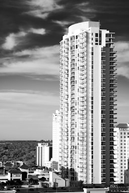 A tall residential skyscraper with numerous balconies stands prominently against a backdrop of a partly cloudy sky. The building features modern architecture and is part of a cityscape with other structures visible in the background, along with some surrounding trees.