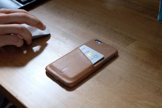 A person's hand is using a computer mouse on a wooden desk. Next to the hand, there is a smartphone with a brown leather case. The case has a slot holding a credit card partially visible.
